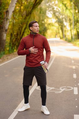 Atletizm kıyafetleri içinde şık bir adam dingin bir ağaçlık yolda duruyor, serin bir güven yayıyor..