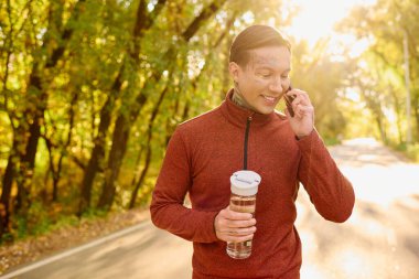Dövmeli adam güneşli bir yolda yürüyor, su içiyor ve telefonda sohbet ediyor..