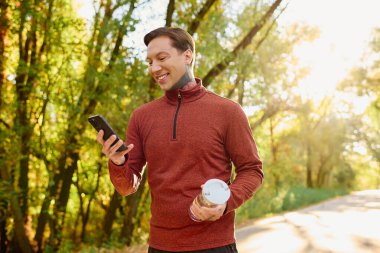 Yakışıklı bir adam güneşli bir parkta geziyor, modaya uygun bir şekilde hava atıyor ve elinde bir su şişesi tutuyor..