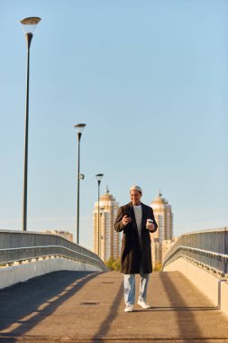 Dövmeleri olan yakışıklı bir adam köprüde duruyor ve güneşin altında kahvesini yudumluyor..