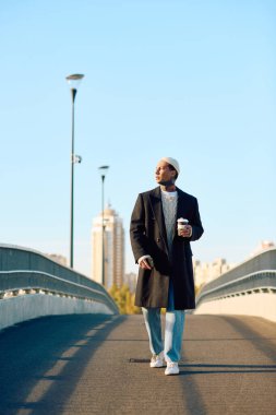 Dövmeli şık bir adam, açık mavi bir gökyüzünün altında, elinde kahve tutarak, sakin bir şekilde yürümekten hoşlanır..