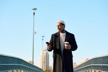 Şık giyinmiş yakışıklı bir adam kahve fincanı tutuyor ve güneşli bir günde telefonunu kullanıyor..