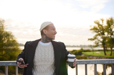 Dövmeleri olan yakışıklı bir adam elinde kahve ve telefon tutarken gülümsüyor..