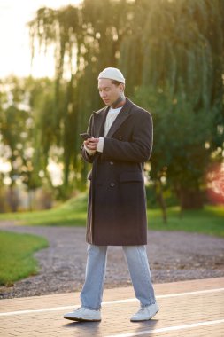 Şık giyinmiş şık bir adam güneşli bir parkta telefonuna odaklanmış duruyor..