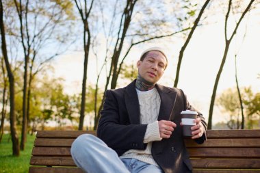 Dövmeleri olan yakışıklı genç adam bankta dinleniyor, sakin bir parkta kahvesini yudumluyor..