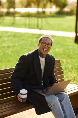 Dövmeli genç adam gülüyor, açık havada yemyeşil bir parkta çalışırken kahvesini yudumluyor..