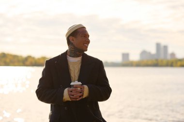 Moda kıyafetli yakışıklı adam, güzel bir günde suyun kenarında gülümserken elinde kahve tutuyor..
