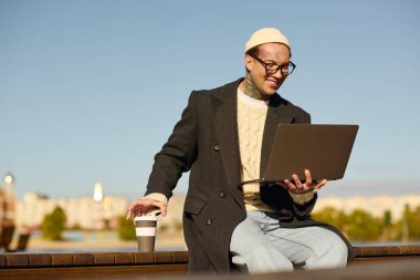 Moda kıyafetleri içinde yakışıklı bir adam laptopuyla dışarıda çalışırken gülümsüyor. Yanında da kahve fincanı var..