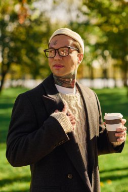 Dövmeleri olan yakışıklı bir adam, sonbaharda canlı bir parkta kahvesini yudumlarken kendine güvenerek poz veriyor..