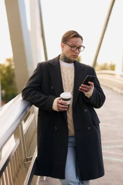 Dövmeli şık bir adam telefonuna bakarken kahvesini yudumluyor..