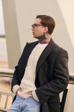 Yakışıklı adam modern bir balkonda kendine güvenerek poz veriyor, moda kıyafetlerini ve dövmelerini sergiliyor..
