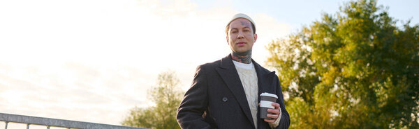 Handsome man with tattoos dressed in modern attire holds a coffee cup while posing outdoors.