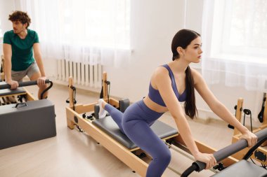 İki birey, iyi aydınlatılmış bir stüdyoda reformist ekipmanlarıyla odaklanmış pilates egzersizleri yapıyor..