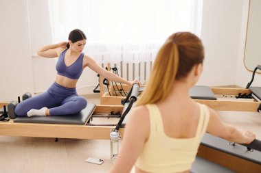 Birlikte egzersiz yapan iki kadın, iyi aydınlatılmış bir stüdyoda pilates rutinlerine odaklanıyorlar..