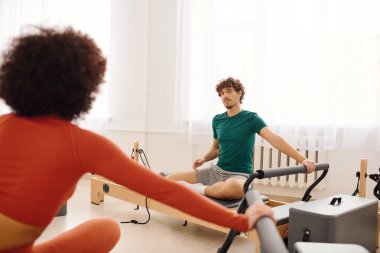 İki insan, parlak ve geniş bir stüdyoda pilates yapıyor. Yeteneklerini ve odaklanmalarını gösteriyorlar..