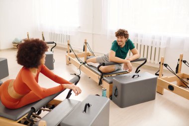 Parlak bir stüdyoda iki kişi, gülümseme ve motivasyon alışverişinde bulunarak pilates çalışmasından zevk alır..