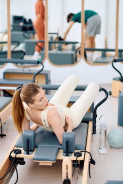 Kendini adamış bir birey, bir reformcu üzerinde pilates yaparken, yakınlarda başkaları pratik yapıyor..