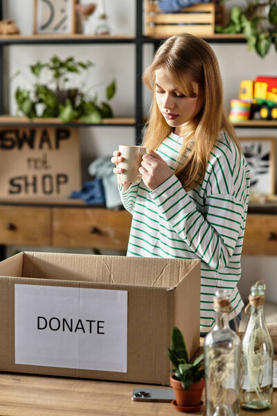 Pretty young woman enjoys a warm drink while sorting clothes for donation in her home environment.