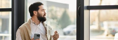 Yakışıklı, sıradan giyinmiş yaşlı bir adam elinde bir kahve fincanı ile dışarı bakıyor..