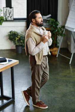 Günlük kıyafetli yaşlı adam, parlak bir çalışma alanında elinde bir fincan kahve tutarken gülümsüyor..