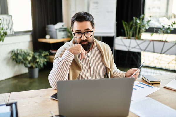 Случайно одетый пожилой человек сосредоточился на ноутбуке, просматривая документы в современном рабочем пространстве.