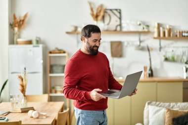 Güzel görünümlü yaşlı bir adam dizüstü bilgisayar tutarken gülümsüyor, rahat ve şık bir oda ile çevrili..