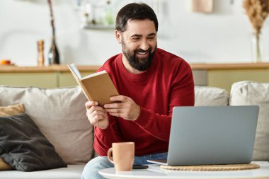 Yaşlı yakışıklı bir adam evinde dizüstü bilgisayarın yanında oturmuş kitap okurken gülümsüyor..