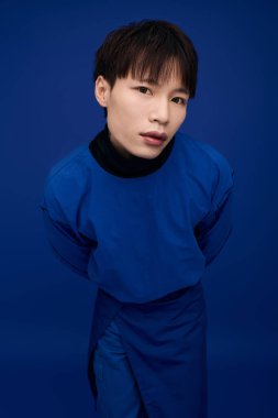 Genç bir Asyalı şık bir şekilde mavi kıyafetler içinde canlı mavi bir arka plana karşı poz veriyor..