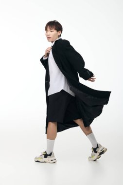 Siyah paltolu ve şortlu kendinden emin genç adam zarif bir şekilde aydınlık bir alanda geziniyor..