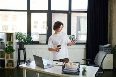 Yakışıklı genç bir adam parlak bir ofiste elinde kahve ve gülümseyerek telefonunu kullanıyor..