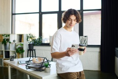 Yakışıklı genç profesyonel şık bir ofiste rahatlıyor, kahvesini yudumluyor ve akıllı telefon kullanıyor..