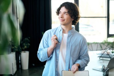 Casual young man thinks deeply with a notebook and pen in a bright office. clipart