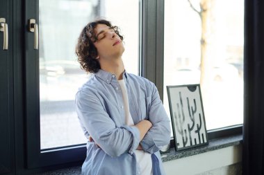 Genç ve çekici bir adam, kollarını pencerenin önünde kavuşturup, ortamı zevkle seyrediyor..