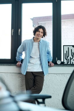 Yakışıklı genç adam pencerenin önünde dikiliyor, modern ofis ortamında kendine güveni tam..