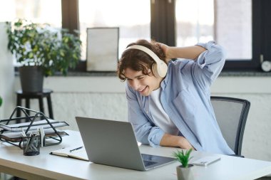 Şık bir ofiste genç bir adam kulaklık takarken dizüstü bilgisayarıyla mutlu bir şekilde etkileşime geçiyor..