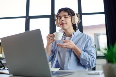 Parlak bir ofiste, genç bir adam kulaklıkla müzik dinlerken sıcak bir içeceğin tadını çıkarıyor..