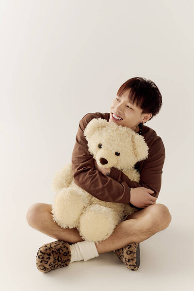 Красивый молодой азиат ярко улыбается, обнимая большого плюшевого мишку в уютной обстановке.