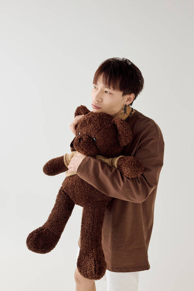 Красивый молодой азиат стоит задумчиво обнимая мягкого плюшевого мишку в ярком пространстве.