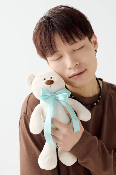 Молодой азиат с закрытыми глазами и улыбкой с любовью держит плюшевого мишку с голубой лентой.