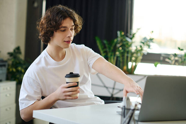 Handsome young professional sits at a desk, sipping coffee and focused on his laptop.
