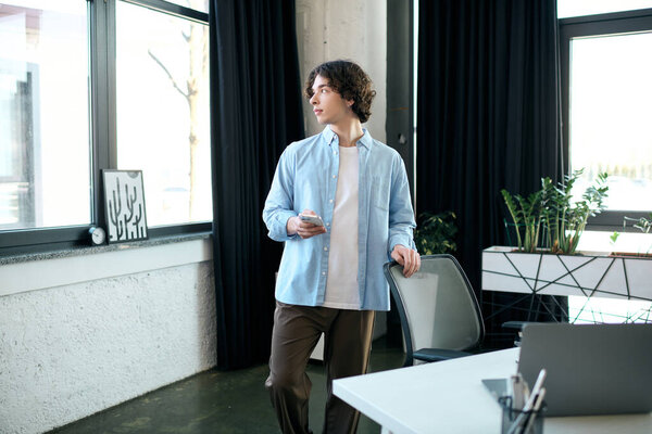 Красивый молодой человек стоит у окна, поглощённый телефоном в стильном офисе.