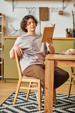 Genç adam rahatça bir sandalyeye oturup, odaklanmış ama rahat bir ifadeyle kitap okuyor..