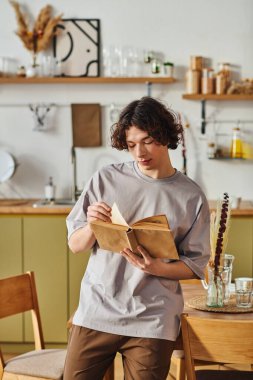 Yakışıklı genç bir adam şık bir mutfakta duruyor, defterine yazmaya odaklanmış..