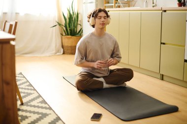 Kulaklıklı genç bir adam güneşli bir ofiste paspasın üzerinde rahatlar, bir yudum içer ve meditasyon yapar..