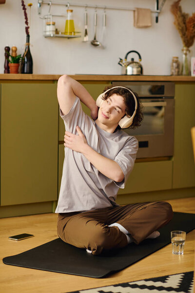 Молодой, привлекательный мужчина сидит скрестив ноги на коврике, растягиваясь и раскручиваясь в своем домашнем офисе.