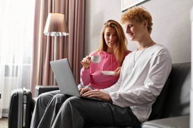 A loving couple relaxes in a stylish hotel room, bonding over coffee and a laptop, sharing joy. clipart