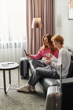 Neşeli bir çift otel koltuğunda oturur, kahvelerini yudumlar ve telefonlarına bakıp gülerler..