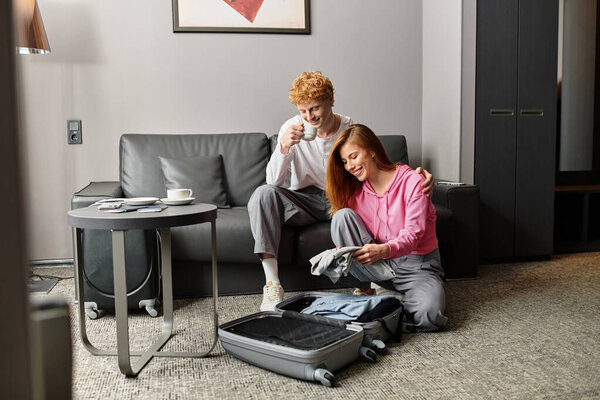 In a cozy hotel room, a couple shares playful moments while unpacking their bags.