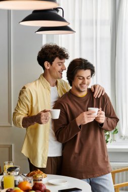 İki erkek, sıcak güneş ışığı altında şık evlerinde kahve içip kucaklaşırlar..