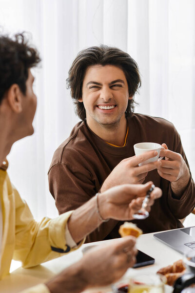 Два красивых мужчины пьют кофе и болтают в своей шикарной квартире, излучая радость.